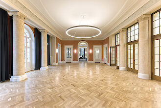 Saal mit historischem Parkettboden, Säulen, großen Fenstern und ringförmiger Pendelleuchte an der Decke.