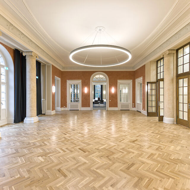 Vestíbulo con parquet histórico, columnas, grandes ventanales y luminaria colgante en forma de anillo en el techo.