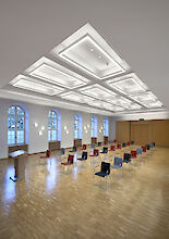 Moderna sala de actos con iluminación geométrica en el techo, grandes ventanales e hileras de sillas frente a un atril.