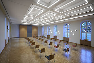 Sala de actos luminosa con ventanas altas, suelo de madera e iluminación geométrica en el techo, asientos para conferencias