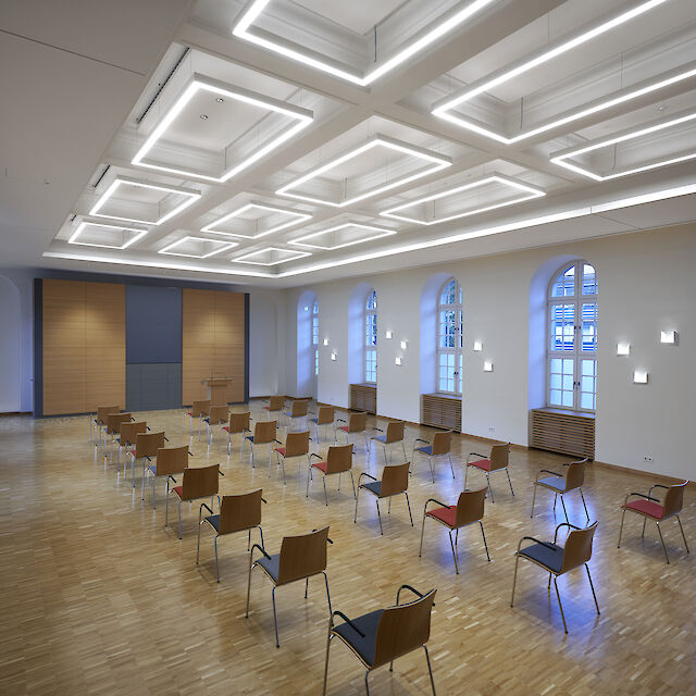Bright event room with high windows, wooden floor and geometric ceiling lighting, seating for lectures