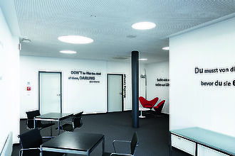 Un moderno salón de reuniones con paredes blancas y mobiliario negro. En el techo hay instaladas luces redondas empotradas que iluminan intensamente la sala. Las paredes están decoradas con citas inspiradoras como 