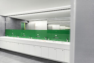 Un cuarto de baño moderno con varios lavabos y tocadores blancos. La pared trasera está revestida de cristal verde, que da una impresión fresca y limpia. Sobre los lavabos hay un gran espejo que amplía visualmente la estancia. El techo está provisto de luces largas y estrechas que garantizan una iluminación uniforme y clara. Las baldosas del suelo son de color gris oscuro y complementan el diseño moderno del cuarto de baño.