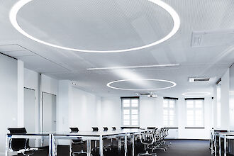 Una moderna sala de reuniones con grandes ventanales y paredes blancas. En el techo hay grandes lámparas redondas que crean una luz uniforme y brillante. La sala está amueblada con largas mesas de reuniones y sillas de oficina negras que crean un ambiente profesional.
