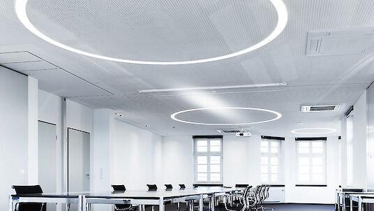 A modern meeting room with large windows and white walls. There are large, round lights on the ceiling that create an even, bright light. The room is furnished with long conference tables and black office chairs that create a professional atmosphere.