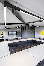 Das Bild zeigt einen modernen Konferenzraum mit einer U-förmigen Tischanordnung. Die Tische sind weiß, und in der Mitte des U befindet sich ein Bereich mit einem grauen Teppichboden. An der Decke hängen schwarze, lineare Lichtstrukturen mit integrierten Spots, die den Raum beleuchten. An der Stirnseite des Raumes ist ein großer Flachbildfernseher auf einem Ständer zu sehen. Links im Bild befindet sich ein Sideboard mit verschiedenen technischen Geräten. Die Wände sind in einem dunklen Grau gehalten.