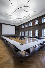 Das Bild zeigt einen großen, modernen Konferenzraum mit hohen Decken. In der Mitte des Raumes steht ein großer U-förmiger Konferenztisch mit weißen Tischplatten und schwarzen Stühlen. Jeder Platz am Tisch ist mit einem Mikrofon ausgestattet. Der Boden ist aus elegantem Holzparkett in einem Fischgrätenmuster. An den Wänden befinden sich hohe Fenster, die viel natürliches Licht hereinlassen. An der Decke hängen mehrere geschwungene, ringförmige Leuchten, die dem Raum ein modernes und stilvolles Aussehen verleihen. An der Stirnseite des Raumes ist eine große Leinwand für Präsentationen installiert.