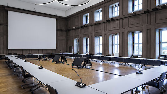 La imagen muestra una sala de conferencias grande y moderna con techos altos. En el centro de la sala hay una gran mesa de conferencias en forma de U con tableros blancos y sillas negras. Cada asiento de la mesa está equipado con un micrófono. El suelo es de elegante parqué de madera en espiga. Las paredes están revestidas de altos ventanales por los que entra mucha luz natural. Del techo cuelgan varias lámparas curvadas en forma de anillo que dan a la sala un aspecto moderno y elegante. En la parte delantera de la sala hay una gran pantalla para presentaciones.