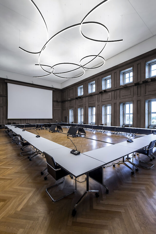 The picture shows a large, modern conference room with high ceilings. In the centre of the room is a large U-shaped conference table with white table tops and black chairs. Each seat at the table is equipped with a microphone. The floor is made of elegant wooden parquet in a herringbone pattern. The walls are lined with high windows that let in plenty of natural light. Several curved, ring-shaped luminaires hang from the ceiling, giving the room a modern and stylish look. A large screen for presentations is installed at the front of the room.