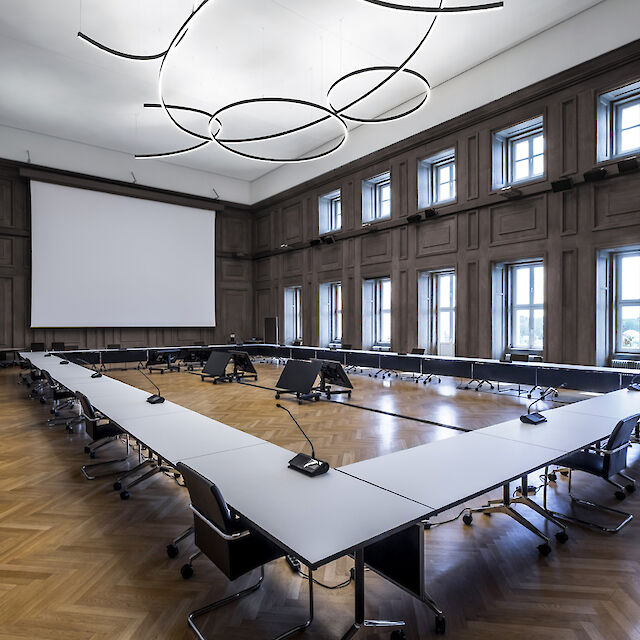 The picture shows a large, modern conference room with high ceilings. In the centre of the room is a large U-shaped conference table with white table tops and black chairs. Each seat at the table is equipped with a microphone. The floor is made of elegant wooden parquet in a herringbone pattern. The walls are lined with high windows that let in plenty of natural light. Several curved, ring-shaped luminaires hang from the ceiling, giving the room a modern and stylish look. A large screen for presentations is installed at the front of the room.