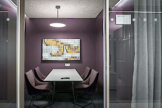 La imagen muestra una pequeña y moderna sala de reuniones con paredes moradas. En el centro de la sala hay una mesa rectangular rodeada por cuatro sillas moradas. Encima de la mesa hay una luminaria colgante redonda y blanca que ilumina la sala. En la pared de detrás de la mesa hay una obra de arte hecha con pequeños azulejos cuadrados de varios colores, principalmente dorados y azules. La habitación está separada del pasillo por una pared de cristal, con una cortina gris colgada en el lado derecho. Sobre la mesa hay un mando a distancia.