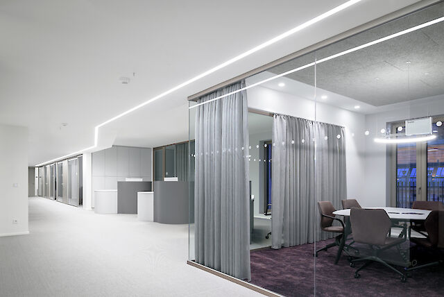 The picture shows a modern office corridor with an open, bright design. On the right-hand side, there is a meeting room and several offices separated from the corridor by a glass wall and grey curtains. The meeting room has a dark purple carpet and is furnished with a round table and several chairs. There is a light line on the ceiling of the corridor that illuminates the entire corridor.