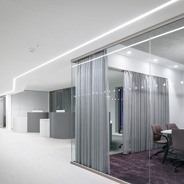 The picture shows a modern office corridor with an open, bright design. On the right-hand side, there is a meeting room and several offices separated from the corridor by a glass wall and grey curtains. The meeting room has a dark purple carpet and is furnished with a round table and several chairs. There is a light line on the ceiling of the corridor that illuminates the entire corridor.