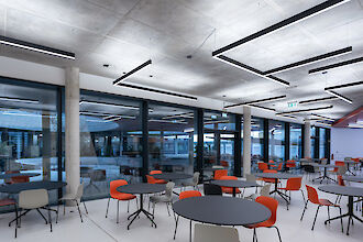 Dieses Bild zeigt einen modernen Cafeteria-Bereich mit Betonwänden und -decke. Von der Decke hängen lange, rechteckige LED-Leuchten in verschiedenen Winkeln. Der Raum ist mit runden Tischen ausgestattet, die jeweils von vier Stühlen umgeben sind. Die Stühle sind in den Farben Orange und Grau gehalten, was dem Raum einen farbenfrohen Akzent verleiht. Auf der linken Seite des Bildes sind große Fenster und Glastüren zu sehen, die den Blick nach draußen ermöglichen und Tageslicht in den Raum lassen. Der Boden ist glatt und weiß, was den modernen und sauberen Look des Raums unterstreicht.