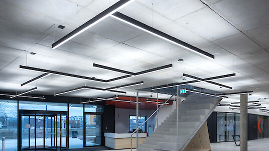 Esta imagen muestra un interior moderno y minimalista con paredes y techo de hormigón. Del techo cuelgan luces LED largas y rectangulares en distintos ángulos, que dan a la habitación un patrón geométrico. En el centro de la sala hay una escalera abierta de hormigón con una barandilla metálica que conduce hacia arriba. A la izquierda hay grandes ventanales y una puerta de cristal que dejan entrar la luz del día. El suelo es liso y blanco, lo que acentúa el aspecto moderno y limpio de la sala.