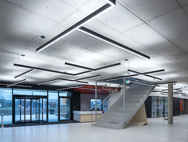This image shows a modern, minimalist interior design with concrete walls and a concrete ceiling. Long, rectangular LED luminaires hang from the ceiling at different angles, giving the room a geometric pattern. In the centre of the room is an open staircase made of concrete with a metal railing that leads upwards. To the left of the picture are large windows and a glass door that let daylight into the room. The floor is smooth and white, which emphasises the modern and clean look of the room.