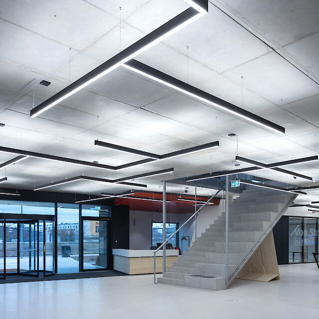 This image shows a modern, minimalist interior design with concrete walls and a concrete ceiling. Long, rectangular LED luminaires hang from the ceiling at different angles, giving the room a geometric pattern. In the centre of the room is an open staircase made of concrete with a metal railing that leads upwards. To the left of the picture are large windows and a glass door that let daylight into the room. The floor is smooth and white, which emphasises the modern and clean look of the room.