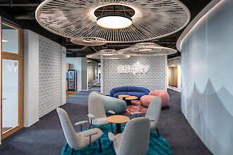 The picture shows a modern office lounge that is stylishly and cosily furnished. In the centre of the room are several upholstered chairs around small round tables on a blue carpet. In the background is a curved, blue and purple couch, accompanied by a pink pouffe. Round pendant luminaires with eye-catching, fan-like design elements hang above the seating. The walls have a grey brick look, with a brick wall in the background bearing the lettering 