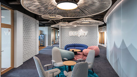 La imagen muestra un moderno salón de oficina amueblado de forma elegante y acogedora. En el centro de la sala hay varias sillas tapizadas alrededor de pequeñas mesas redondas sobre una alfombra azul. Al fondo hay un sofá curvo, azul y morado, acompañado de un puf rosa. Sobre los asientos cuelgan lámparas colgantes redondas con llamativos elementos de diseño en forma de abanico. Las paredes tienen un aspecto de ladrillo gris, con una pared de ladrillo al fondo con la inscripción 