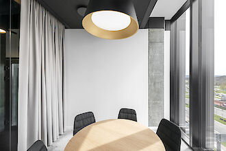The picture shows a modern, minimalist meeting room. In the centre is a round wooden table surrounded by four black, upholstered chairs. A large, round pendant luminaire with a black exterior and a golden interior hangs from the ceiling, illuminating the room. On the left is a large, white curtain covering a door. On the right, there is a large window that lets in daylight and offers a view of a cityscape. The walls are plain white and grey, giving the room an elegant and uncluttered look.