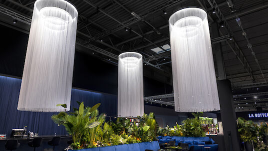 La imagen muestra un interior moderno y elegante con tres grandes luminarias cilíndricas que cuelgan del techo. Cada luminaria está formada por muchos hilos finos que crean un aspecto impresionante, casi fluido. Debajo de las luces hay exuberantes arreglos vegetales que aportan verdor a la habitación. Al fondo se ve un largo banco azul y varias sillas negras. El techo es alto y de diseño industrial, lo que da a la sala una sensación de amplitud y ventilación.