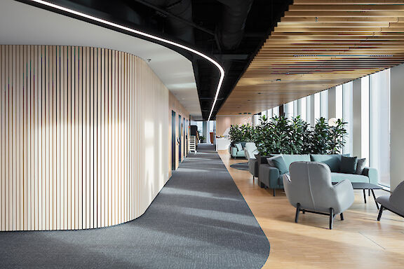 Modern office corridor with curved light line, wooden slat ceiling and lounge area with armchairs.