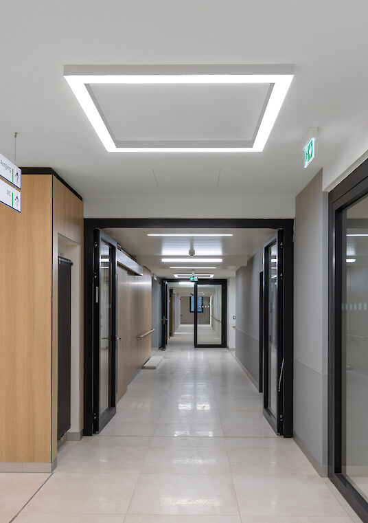 Pasillo luminoso con iluminación cuadrada «Matric» en el techo, puertas y baldosas claras en el suelo.