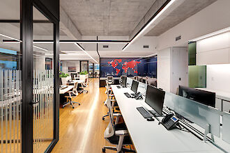 A modern office with an open-plan design. The floor is made of light-coloured wood and the ceiling has a raw concrete look. In the centre of the room are long desks with white table tops and modern office chairs. There are several computer screens and telephones on the desks. Along the left-hand side of the picture there is a glass wall with a door leading into another office space. On the back wall of the office is a world map in red, applied to a dark, glossy surface. Modern light lines run above the work areas and are integrated into the ceiling design, providing bright and even lighting.