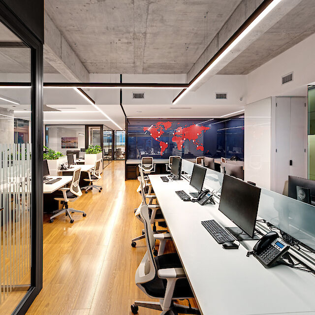 A modern office with an open-plan design. The floor is made of light-coloured wood and the ceiling has a raw concrete look. In the centre of the room are long desks with white table tops and modern office chairs. There are several computer screens and telephones on the desks. Along the left-hand side of the picture there is a glass wall with a door leading into another office space. On the back wall of the office is a world map in red, applied to a dark, glossy surface. Modern light lines run above the work areas and are integrated into the ceiling design, providing bright and even lighting.
