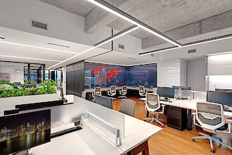 A modern office with an open-plan design. The floor is made of light-coloured wood and the ceiling has a raw concrete look. In the centre of the room are long desks with white table tops and modern office chairs. There are several computer screens and telephones on the desks. On the back wall of the office is a world map in red, applied to a dark, glossy surface. Modern light lines run above the work areas and are integrated into the ceiling design, providing bright and even lighting.