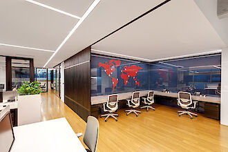 A modern office with an open-plan design. The floor is made of light-coloured wood and the ceiling has a raw concrete look. In the centre of the room are long desks with white table tops and modern office chairs. There are several computer screens and telephones on the desks. On the back wall of the office is a world map in red, applied to a dark, glossy surface. Modern light lines run above the work areas and are integrated into the ceiling design, providing bright and even lighting.