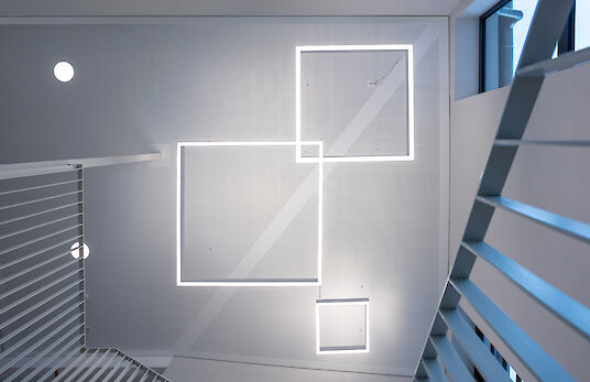 Upward view of square Matric pendant luminaires in a staggered arrangement between two flights of stairs.