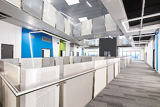 Vista interior del Instituto Chisholm de Melbourne, Australia. La foto muestra un luminoso pasillo con líneas de luz modulares de la línea de productos Matric de Lightnet instaladas en el techo. La sala cuenta con barandillas metálicas y paredes de cristal integradas en un diseño minimalista. Los grandes ventanales del fondo dejan entrar la luz del día y acentúan la arquitectura espaciosa y abierta.
