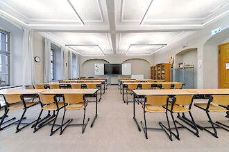 Das Bild zeigt einen hellen, modernen Klassenraum. In der Mitte des Raums stehen mehrere Reihen von Tischen und Stühlen, die aus hellem Holz und schwarzem Metall bestehen und ordentlich in Reihen angeordnet sind. Die Decke ist weiß, mit rechteckigen Pendelleuchten. An der Rückwand des Raums befindet sich ein Whiteboard und ein großer Bildschirm. Rechts stehen zwei Aktenschränke und ein offenes Regal mit Ordnern und Büchern. Links sind große Fenster mit weißen Vorhängen, die viel Tageslicht hereinlassen.