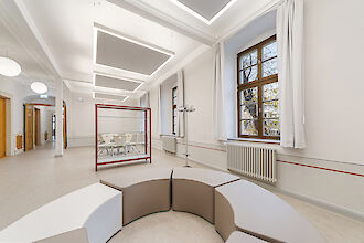 Das Bild zeigt einen modernen Innenbereich mit einem großen, hellen Raum. Im Vordergrund befindet sich eine runde, modulare Sitzgelegenheit mit beige und weißen Elementen. Im Hintergrund sind große Fenster mit weißen Vorhängen zu sehen, durch die Tageslicht hereinfällt. Rechts an der Wand steht ein Heizkörper, und links im Raum befindet sich ein offener Durchgang zu einem weiteren Bereich. An der Decke hängen moderne rechteckige Leuchten mit Akustikelement. In der Mitte des Raumes steht eine Glasvitrine mit rotem Metallrahmen.