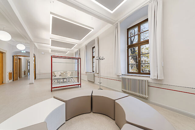 The picture shows a modern interior with a large, bright room. In the foreground is a round, modular seating area with beige and white elements. Large windows with white curtains can be seen in the background, allowing daylight to flood in. There is a radiator on the right-hand wall and an open passageway to another area on the left-hand side of the room. Modern rectangular luminaires with acoustic elements hang from the ceiling. In the centre of the room is a glass cabinet with a red metal frame.


