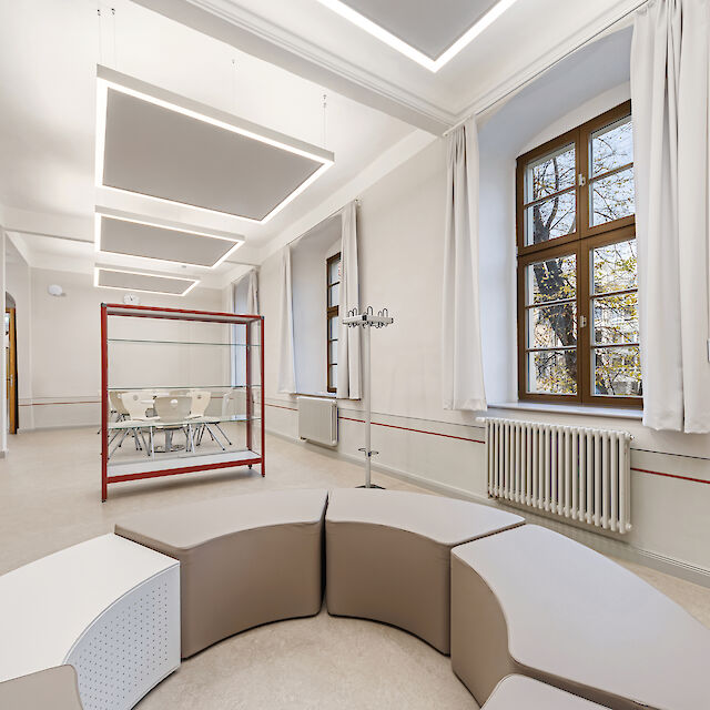 Das Bild zeigt einen modernen Innenbereich mit einem großen, hellen Raum. Im Vordergrund befindet sich eine runde, modulare Sitzgelegenheit mit beige und weißen Elementen. Im Hintergrund sind große Fenster mit weißen Vorhängen zu sehen, durch die Tageslicht hereinfällt. Rechts an der Wand steht ein Heizkörper, und links im Raum befindet sich ein offener Durchgang zu einem weiteren Bereich. An der Decke hängen moderne rechteckige Leuchten mit Akustikelement. In der Mitte des Raumes steht eine Glasvitrine mit rotem Metallrahmen.