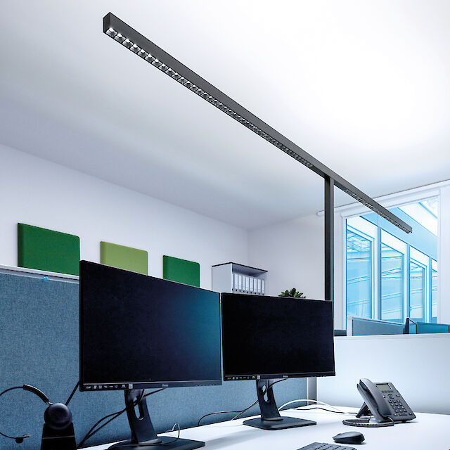 The picture shows a modern office with a long, narrow, black free-standing luminaire. Under the lamp are two large computer screens on a white desk, next to a telephone and a keyboard. In the background, green and grey acoustic panels are mounted on the wall and a window lets in daylight.