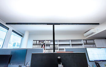 Entorno de oficina moderno con una lámpara de pie larga y delgada que se extiende por el centro de varios escritorios. Las mesas están equipadas con monitores y material de oficina. Al fondo se ven grandes ventanales y estanterías con archivadores. En la pared hay un aparato de aire acondicionado.