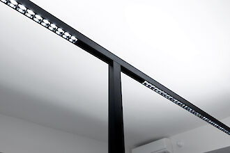 Primer plano de una esbelta lámpara de pie negra de Lightnet. La luminaria está encendida y muestra las fuentes de luz LED dispuestas con precisión. Al fondo se ve parte de un techo blanco y una unidad de aire acondicionado en la pared.