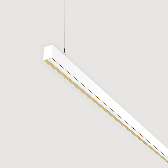 The picture shows a section of a linear white pendant light with a recessed smooth cover. The underside has a gold-coloured reflector ring. The luminaire has a modern design and is suspended from the ceiling on a thin cable. The background is grey.