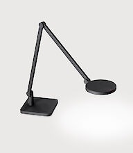 Das Bild zeigt eine moderne, minimalistische Tischleuchte. Die Leuchte ist schwarz und hat einen schlanken, beweglichen Arm, der in zwei Segmente unterteilt ist. Der Leuchtenkopf ist rund und flach. Die Basis der Leuchte ist quadratisch. Die Leuchte steht vor einem hellgrauen Hintergrund.