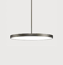 The picture shows a modern, round pendant luminaire with a flat, grey housing. The luminaire has a minimalist design and hangs from the ceiling on a cable. The background colour of the picture is a light grey.