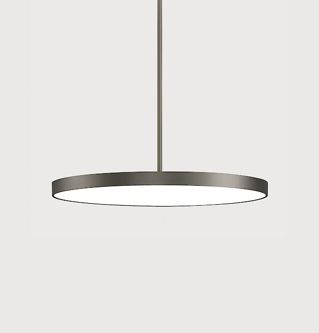 The picture shows a modern, round pendant luminaire with a flat, grey housing. The luminaire has a minimalist design and hangs from the ceiling on a cable. The background colour of the picture is a light grey.
