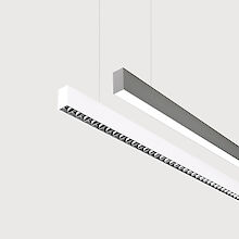 The picture shows a section of two linear pendant luminaires, one with a white and one with a grey surface. The underside of the white luminaire is fitted with a silver-coloured lenticular louvre, with each lens containing a small LED light. The grey luminaire has a smooth cover. Both luminaires have a modern design and are suspended from the ceiling on thin cables. The background is grey.