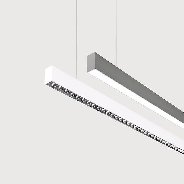 The picture shows a section of two linear pendant luminaires, one with a white and one with a grey surface. The underside of the white luminaire is fitted with a silver-coloured lenticular louvre, with each lens containing a small LED light. The grey luminaire has a smooth cover. Both luminaires have a modern design and are suspended from the ceiling on thin cables. The background is grey.