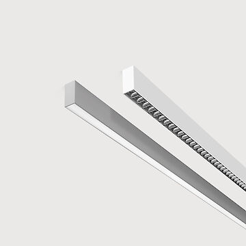 The picture shows a section of two linear surface-mounted luminaires, one with a white and one with a grey surface. The underside of the white luminaire is fitted with a black louvre, with each lens containing a small LED light. The grey luminaire has a smooth cover. Both lights have a modern design. The background is grey.