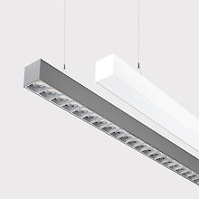 The picture shows a section of two linear pendant luminaires, one with a white and one with a grey surface. The underside of the grey luminaire is fitted with a silver-coloured lenticular louvre, with each lens containing a small LED light. The white luminaire has a smooth cover. Both luminaires have a modern design and are suspended from the ceiling on thin cables. The background is grey.