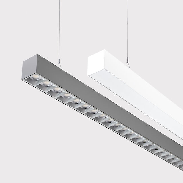 The picture shows a section of two linear pendant luminaires, one with a white and one with a grey surface. The underside of the grey luminaire is fitted with a silver-coloured lenticular louvre, with each lens containing a small LED light. The white luminaire has a smooth cover. Both luminaires have a modern design and are suspended from the ceiling on thin cables. The background is grey.