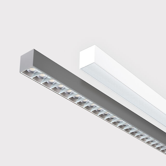 The picture shows a section of two linear surface-mounted luminaires, one with a white and one with a grey surface. The underside of the grey luminaire is fitted with a silver-coloured louvre, with each lens containing a small LED light. The white luminaire has a smooth cover. Both luminaires have a modern design. The background is grey.